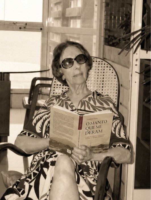 Leonor Rato sentada na cadeira de balanço, lendo o livro O Manto que me Deram